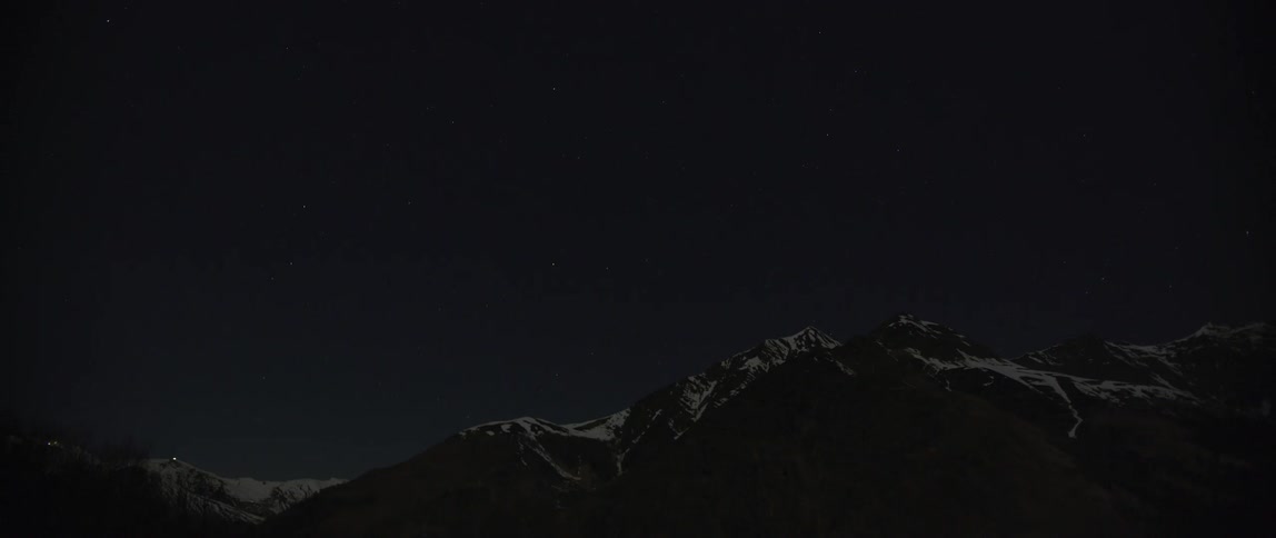 A distance from large craggy mountain lights moves along snowy slope at night