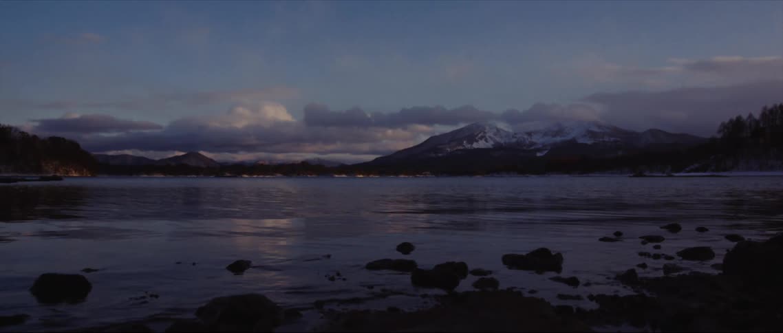 Waves gently splash against rocks as they roll into shore and there are mountains in the distance under a cloudy sky