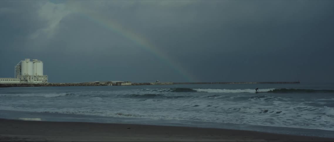 Surfers surf on the ocean under a rainbow on a cloudy day