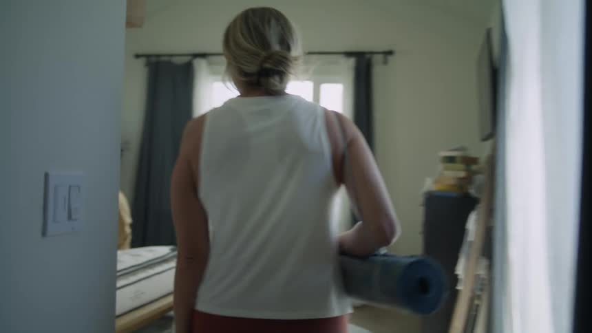 A woman carries her yoga mat in to her bedroom where her son who is on her bed with his dad and brother laughs as he hits her with a pillow
