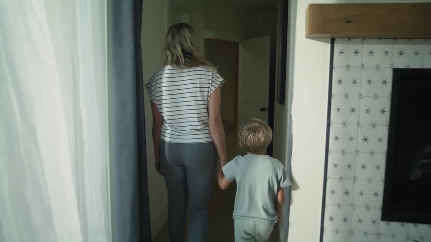 A woman leads a boy by the hand through a corridor in a house during the day