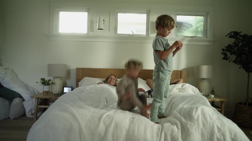 Mom and dad are lying in bed as their two young boys jump up and down on the mattress