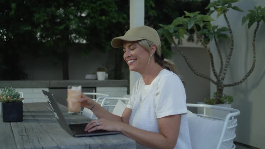 A female wearing a hat is smiling and drinking by her laptop
