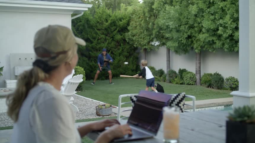 A mom on her laptop watching her husband and son play baseball in the yard