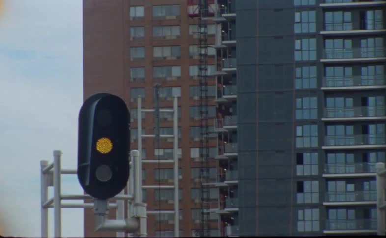 A yellow streetlight flashes on a city street with high rise apartments in the distance