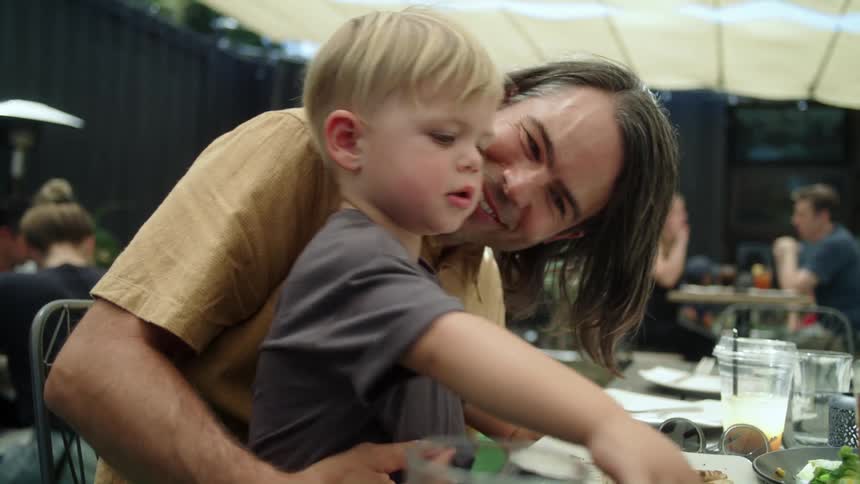 A little boy sits on his dads knee in a cafeteria as his dad talks to him