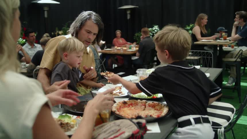 A woman sits next to her son while he hands a pizza slice to his younger brother to eat