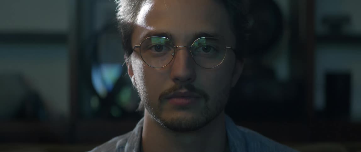 A man sitting and staring straight ahead with a color display reflected on his eyeglasses