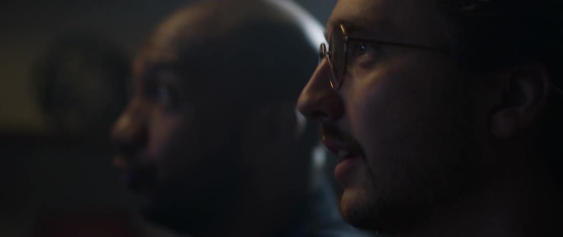 Two men are watching something on tv and something surprises them that makes them start laughing