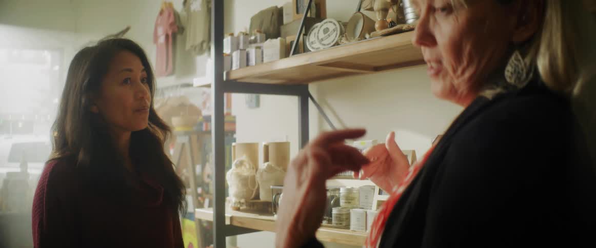 A pair of women talk with one another while standing near a shelf in a shop indoors during the day