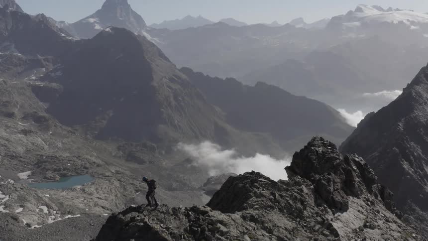 A person is standing high on a mountain ridge looking down at a lake and more mountains in the distance