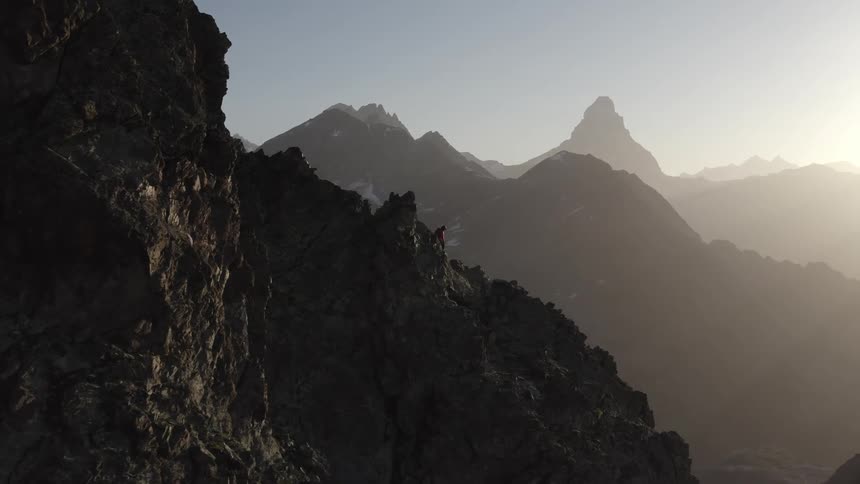 A mountain climber is standing on a mountain ridge getting their rope ready to climb down and there are mountains in the distance in a hazy sky