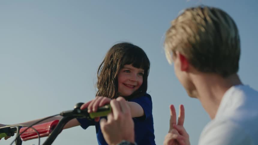 A little girl is sitting on her bicycle holding the handlebars and smiling as she talks to her father