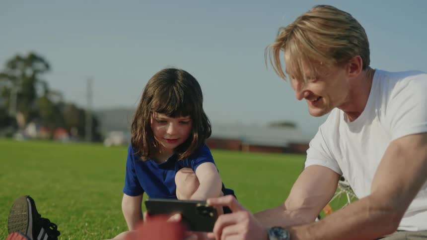 Father and daughter sit on lawn sharing smartphone