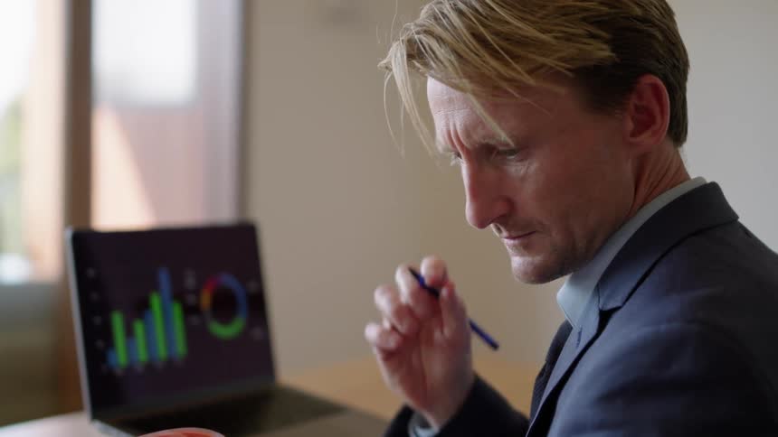 A man in a suit is sitting at a desk with a laptop computer and holding a pen as he looks though a book