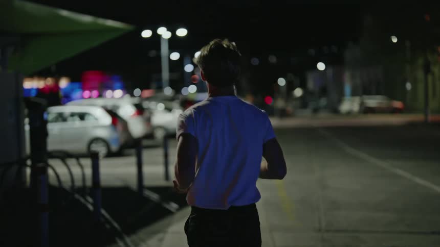 Blonde man wearing white t shirt and black shorts runs by building and cars in car park