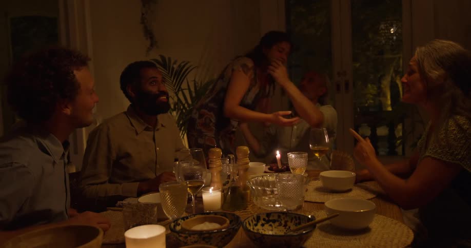 Woman brings small birthday cake to middle aged man sitting at dining able with his wife and friends in the room at night