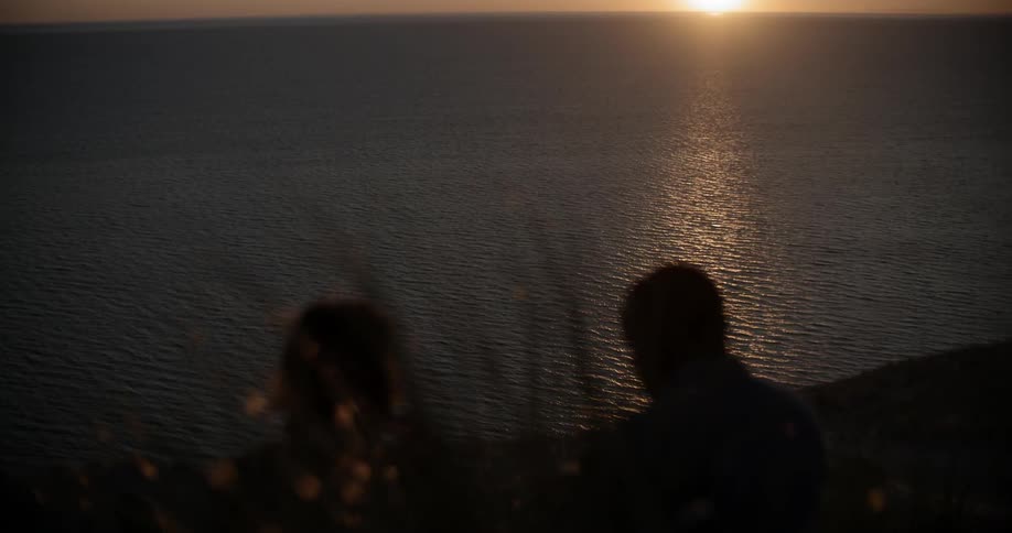 Two people sit on a hill by the water at dawn