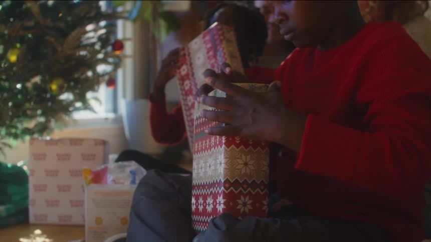 Children prepare to open presents while sitting on the floor with a pair of adults in front of a christmas tree