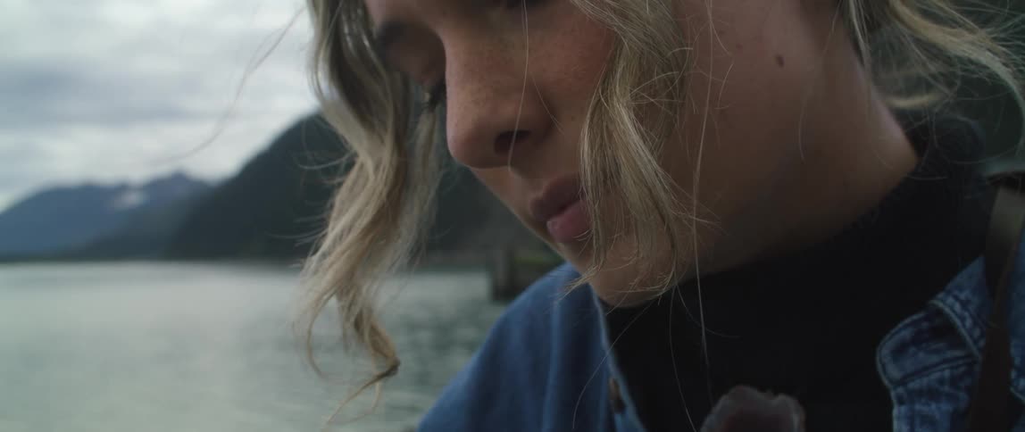 Wind moves womans hair while she stands with misty mountains on shore