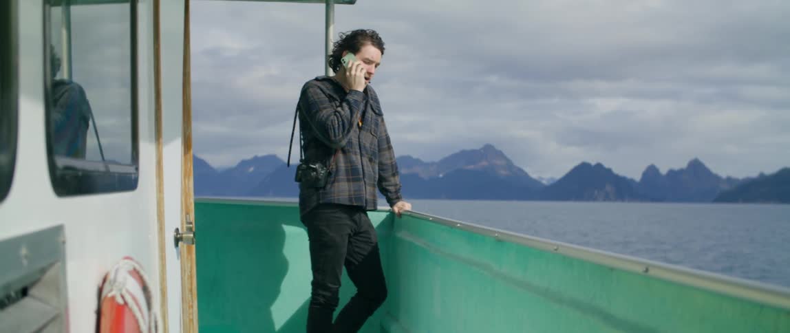 Man carrying camera steps out to lean on side of boat while talking on smartphone as mist covers mountain on shore