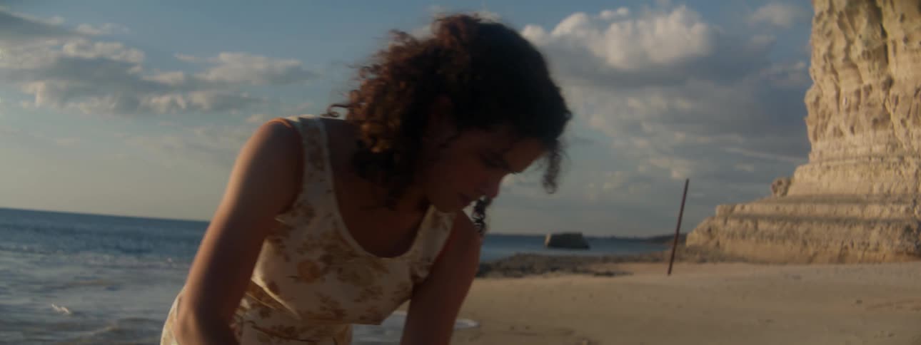 Woman with curly hair and floral dress leans over on the beach near the ocean