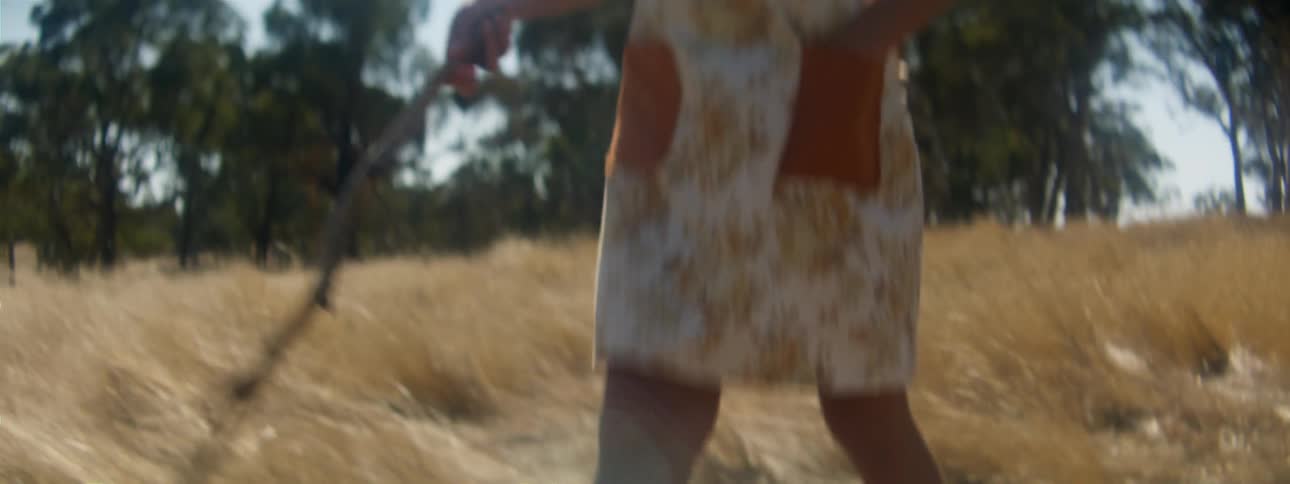 Woman in floral dress walks along brown grassland area holding a stick