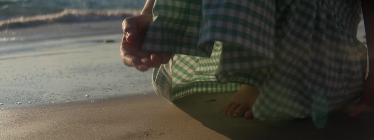 A barefoot girl picking up a seashell while walking on a sandy beach