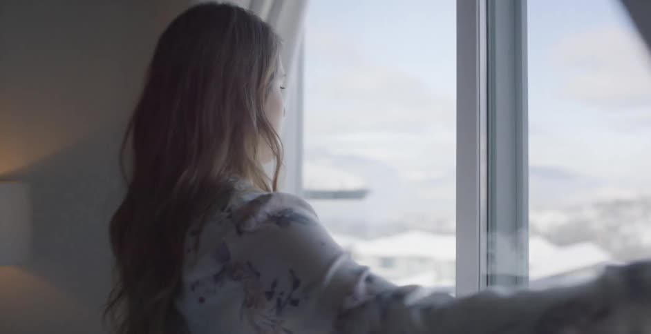 Woman in bedroom opens curtains to look out at city covered in snow