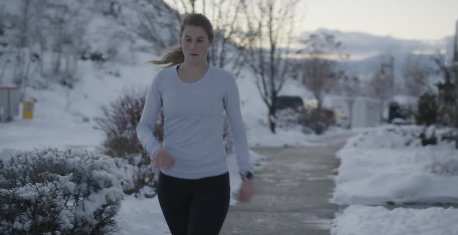 Woman with ponytail white sweatshirt and leggings jogs on the road next to buildings checking her smartwatch in the snowy neighborhood