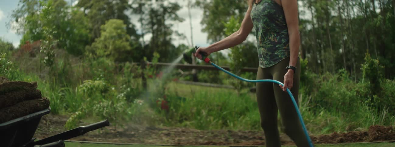 Woman with hat green top and pants hose the lawn in the backyard