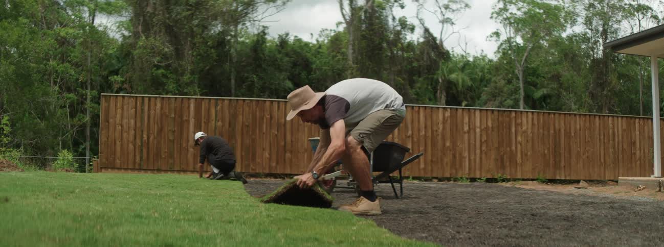 Two men in hat t shirt and shorts lay sod in the garden