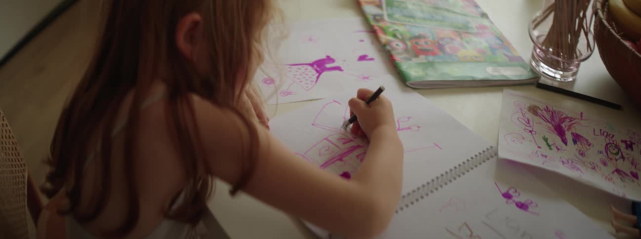 A young girl draws in a sketchbook using a purple crayon