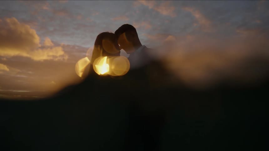 Groom in suit and bride in wedding dress holding hands lovingly looking into each others eyes in sea at sunset