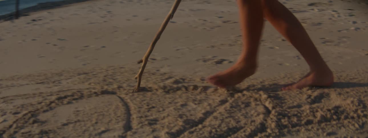 A girl in a dress is walking barefoot on the beach as she draws in the sand with a stick