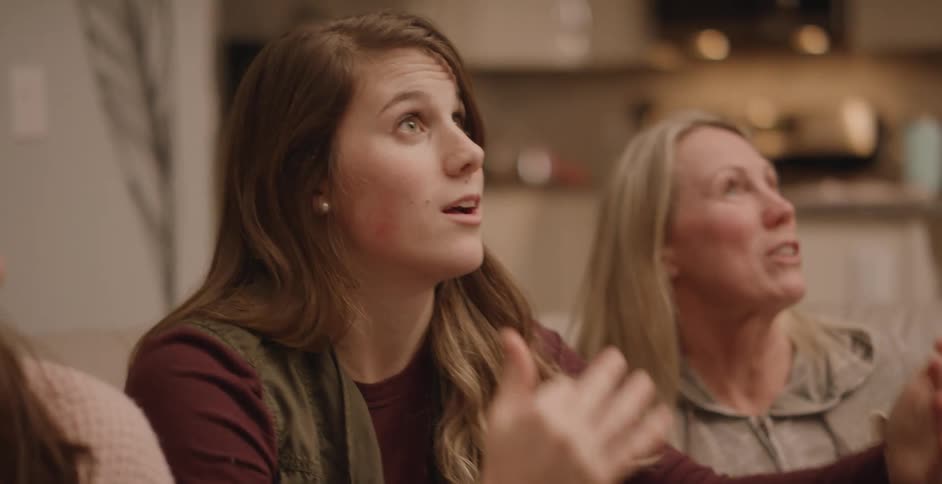Middle aged blonde woman and girls with brown wavy hair sit at table looking up and talking in the kitchen