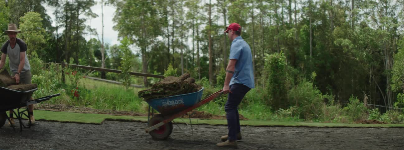 A man in a red hat pushing a wheelbarrow over to another man who is placing grass down