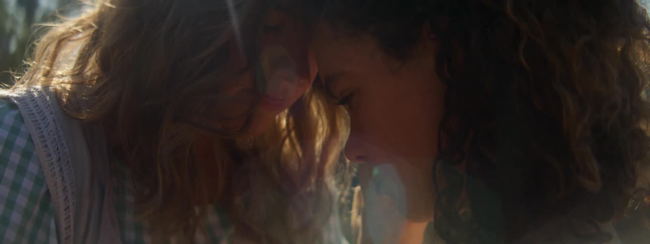 One woman touches anothers cheek as they lean their foreheads together outdoors on a sunny day