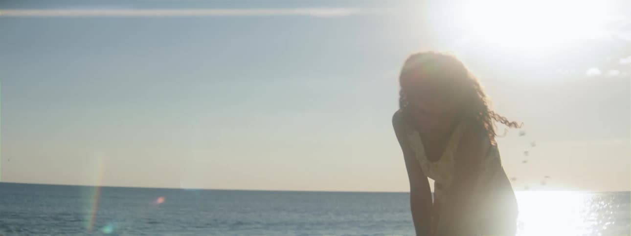 A girl in a dress is walking barefoot on the beach as she draws a line in the sand with a stick