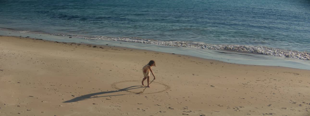 A young woman uses a stick to draw a circle around her in the sand on a beach during the day