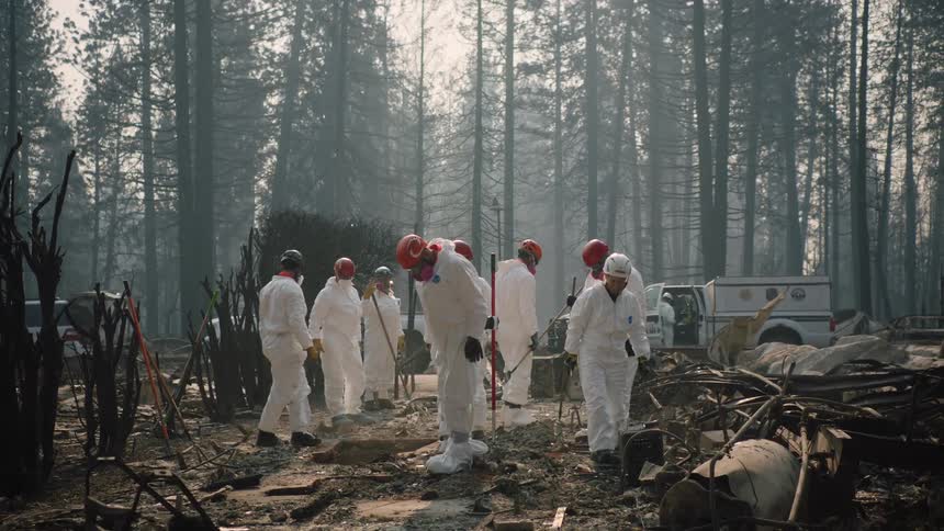 An emergency response team in white jumpsuits are investigating the debris where a building once stood and smoke lingers in the air