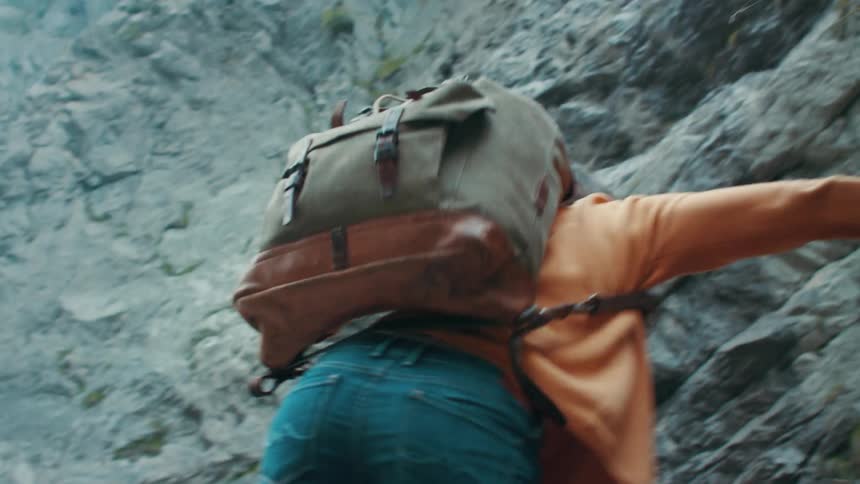 Woman with brown hair orange sweater jeans and backpack climbs up steep mountain