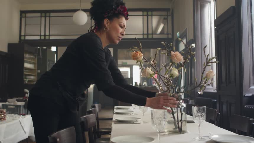 A woman is standing over a table in a restaurant fixing the flowers