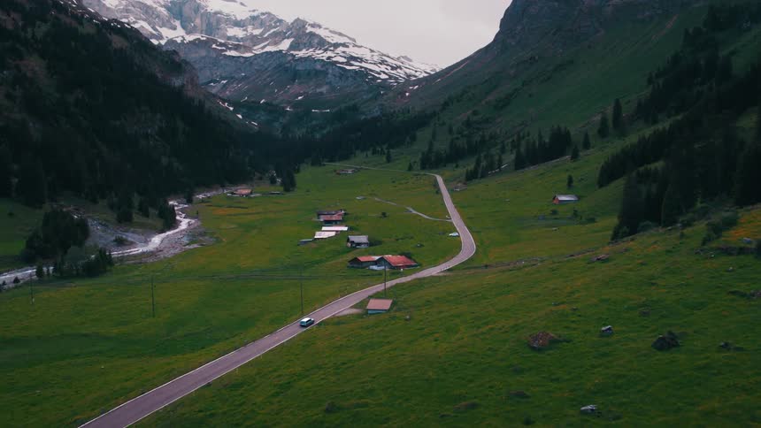 A vehicle drives down a narrow road below a mountain range