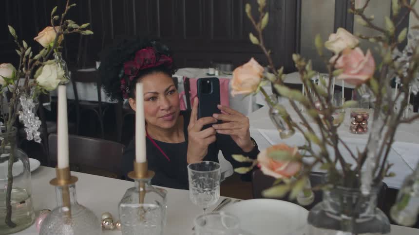 A woman uses her smartphone to photograph flowers on a large dining table