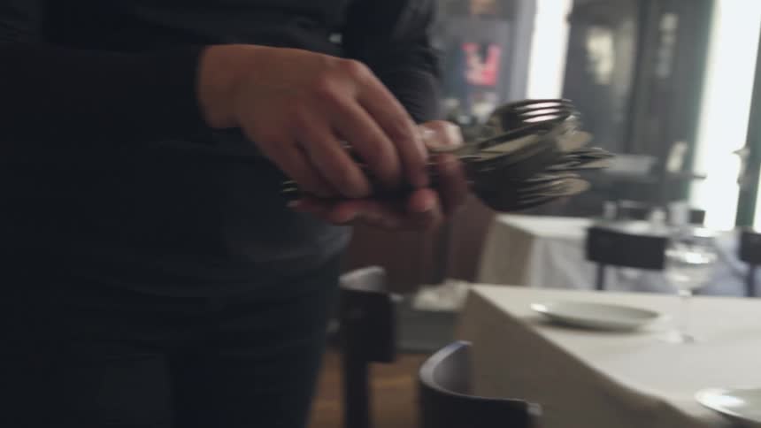 A man is standing in a restaurant holding silverware and setting tables