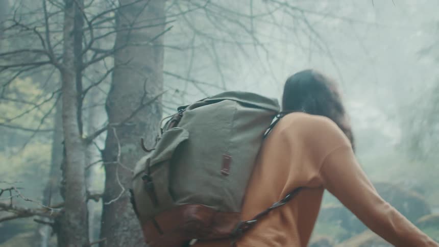 Woman with wavy hair orange sweater backpack and jeans hikes in the foggy woods