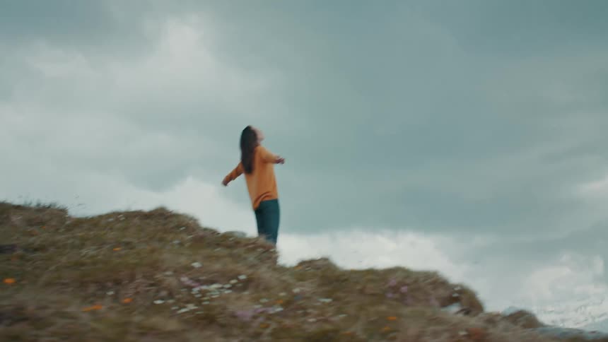 Woman with orange sweater and jeans stands at the edge of high mountain stretching arms on cloudy day