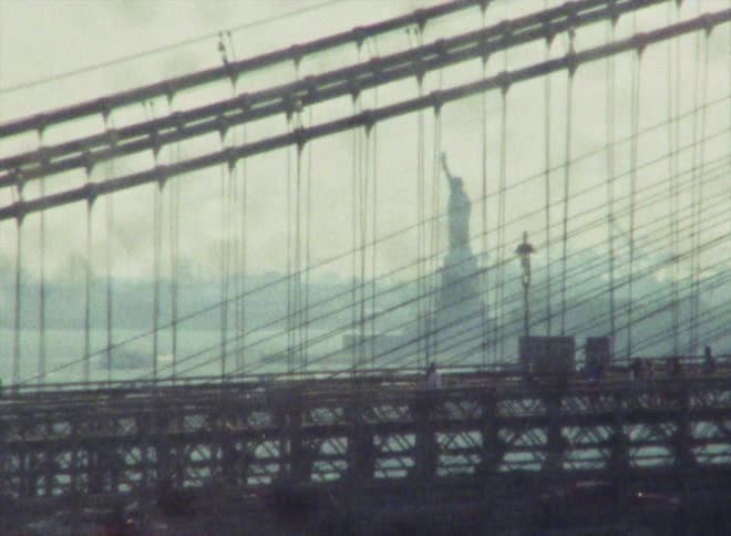 Vehicles pass by people walking down a parallel section on a suspension bridge
