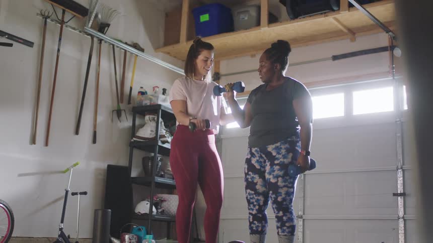 Woman with brown hair pink t shirt and red yoga pants demonstrates how to lift dumbbells to woman with bun green t shirt and floral yoga pants in room